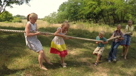 Group of happy school kids playing tug-of-war in city park with teacher.