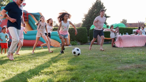 Slow Motion Shot Of Children Playing Football Match With Adults At Summer Garden