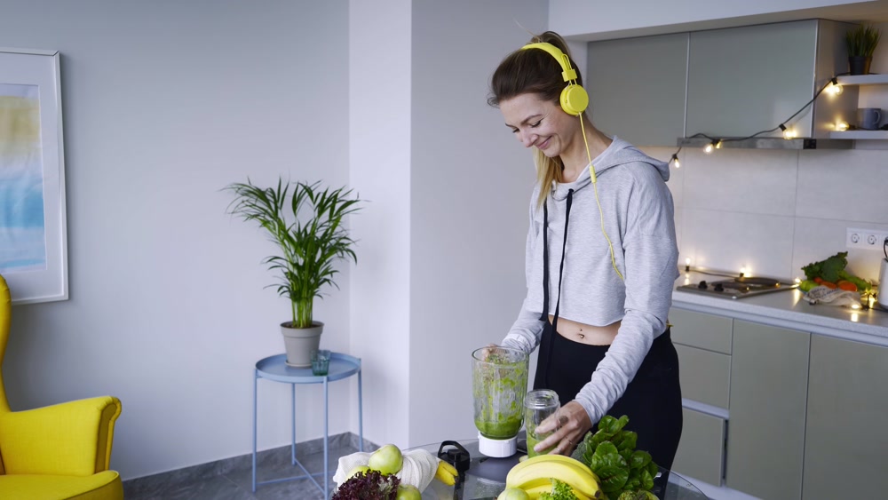 Lady with headphones pours smoothie into bottle in kitchen Stock