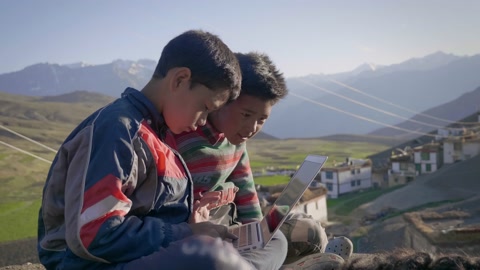 two East Asian rural kids sitting together outdoors engrossed in laptop