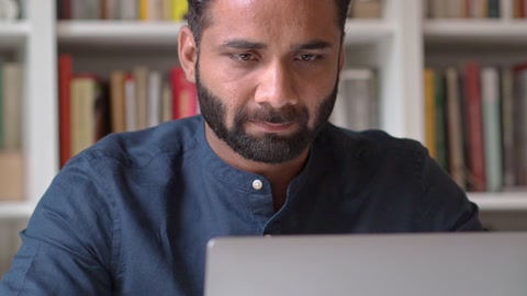 Indian business man working on laptop computer thinking sitting at home office.